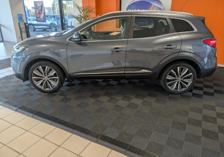 Profil côté gauche d'un Renault Kadjar gris 2017 exposé en intérieur, avec jantes bi-ton et toit ouvrant visible.