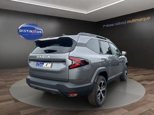 Vue 3/4 arrière droite du Dacia Bigster gris foncé avec feux arrière LED et barres de toit noires dans un showroom.