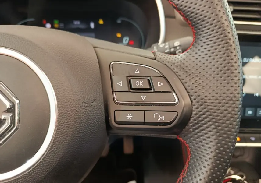Gros plan sur le volant noir perforé avec surpiqûres rouges du MG ZS Luxury 2024, boutons de commande multifonctions visibles.
