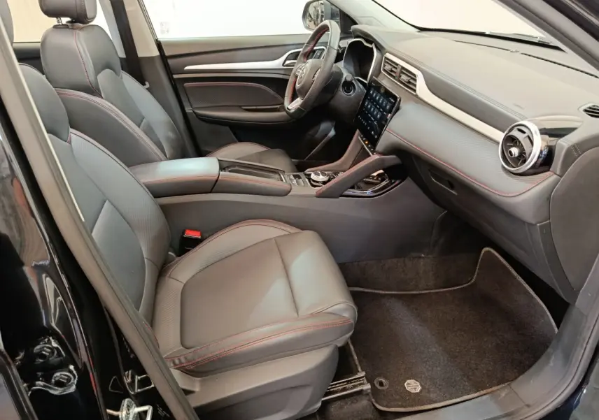 Vue intérieure côté conducteur du MG Motor ZS Luxury 70kWh 2024 noir, sièges cuir avec surpiqûres rouges et écran tactile central.