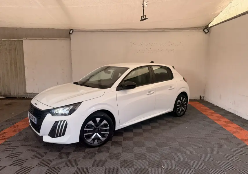 Peugeot 208 blanche vue en 3/4 avant droit, avec jantes noires et phares LED allumés, stationnée en intérieur.