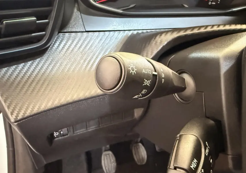 Gros plan sur la commande des phares et le volant noir avec insert carbone du tableau de bord de la Peugeot 208 blanche.