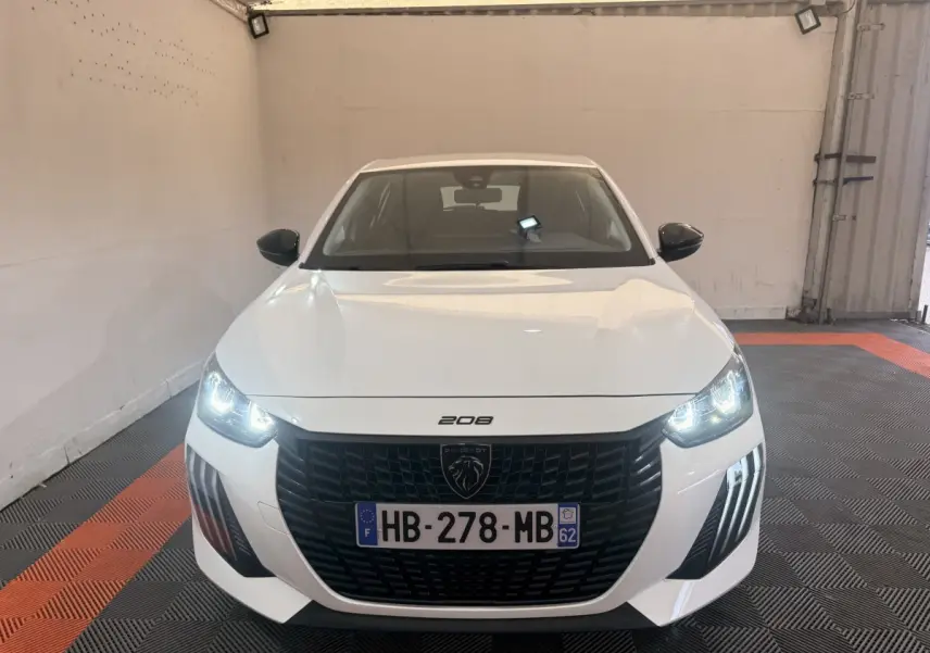 Vue de face d'une Peugeot 208 blanche avec phares allumés et calandre noire dans un garage.