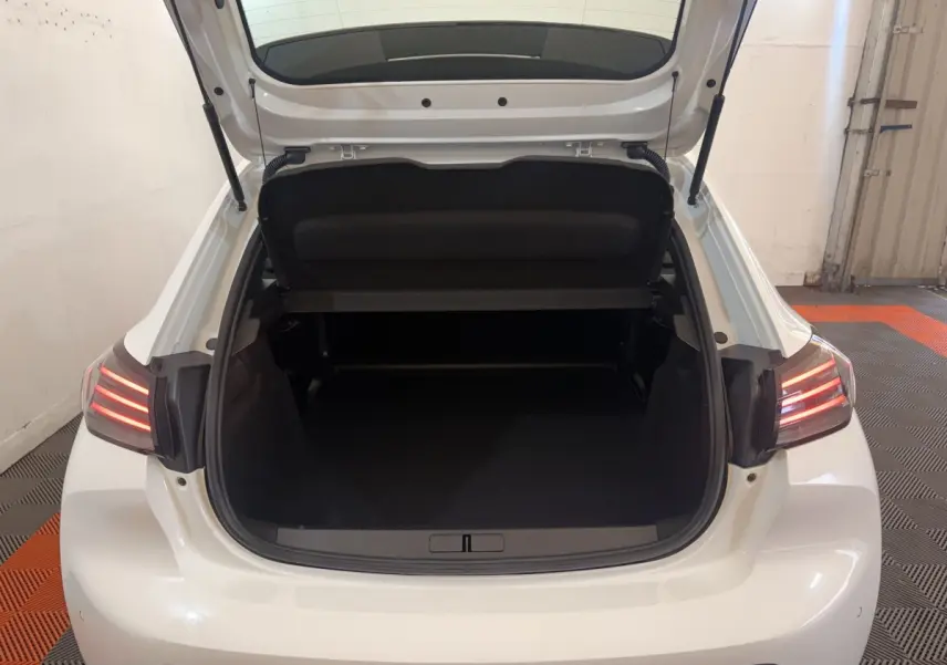 Vue arrière ouverte du coffre d'une Peugeot 208 blanche 2025 avec feux arrière LED allumés dans un garage.