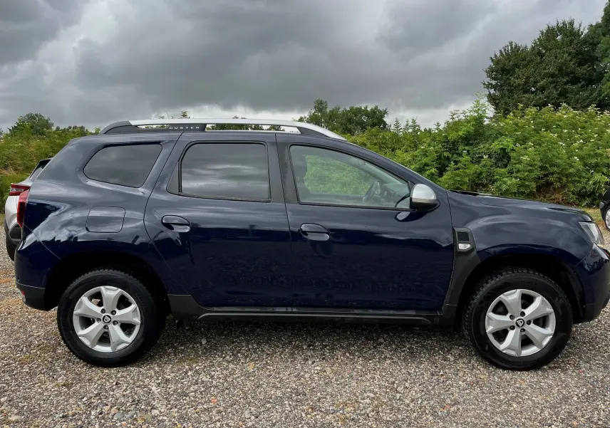 Profil côté gauche d'un Dacia Duster 1.5 DCI bleu marine 2018 avec barres de toit argentées sur un sol gravillonné.
