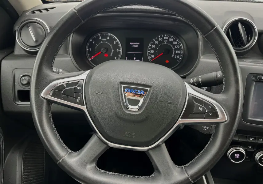 Volant noir de Dacia Duster 1.5 DCI 2018 avec commandes intégrées, tableau de bord et compteurs visibles en arrière-plan.