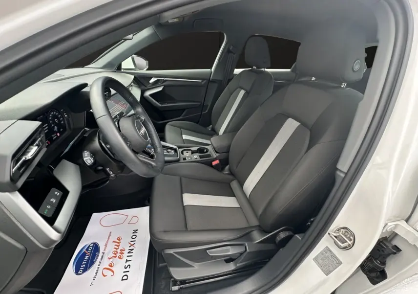 Vue intérieure côté conducteur de l'Audi A3 blanc 2024, sièges tissu noir avec bandes grises et tableau de bord moderne.