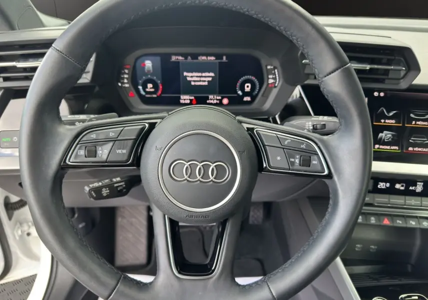 Vue centrée sur le volant noir de l'Audi A3 2024 blanc, avec tableau de bord numérique et commandes intégrées visibles.