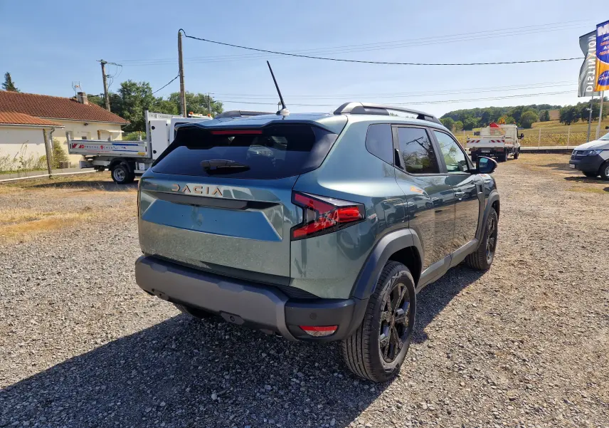 Vue 3/4 arrière droite du Dacia Duster 1.8 Hybrid 155 Extreme vert cèdre avec jantes noires et toit équipé de barres de toit.