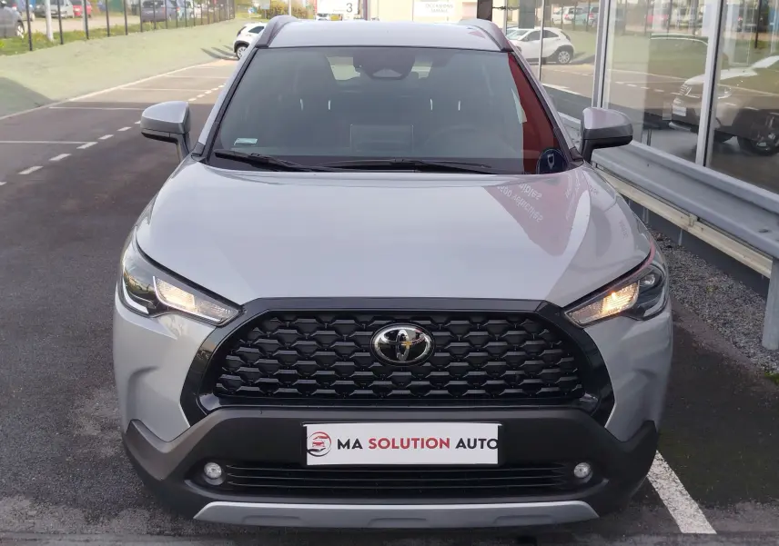 Vue frontale d'un Toyota Corolla Cross 2025 gris acier avec calandre noire et feux allumés sur un parking.