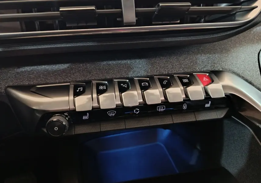Gros plan sur la rangée de boutons argentés et noirs du tableau de bord du Peugeot 3008 blanc, avec éclairage bleu dessous.