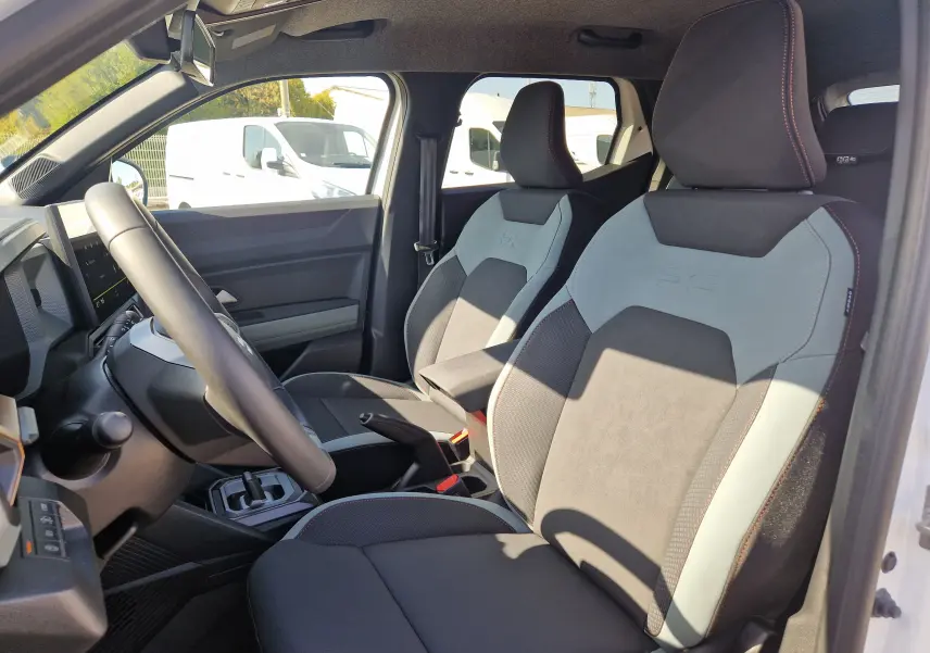 Vue intérieure côté conducteur du Dacia Duster blanc 2026, mettant en valeur les sièges bi-ton gris et noir avec surpiqûres rouges.