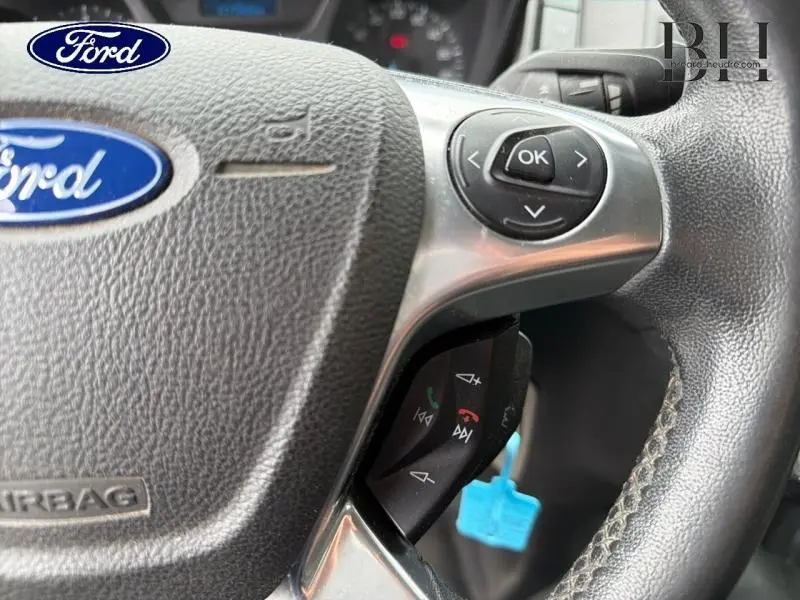 Gros plan sur le volant noir du Ford Transit Custom avec commandes audio et logo Ford visible au centre.