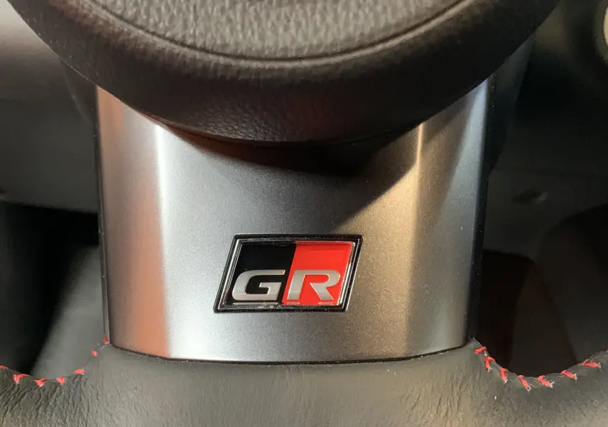 Gros plan sur le logo GR rouge et noir sur le volant en cuir noir avec surpiqûres rouges de la Toyota Yaris 1.6 VVT-I 261 Premium.