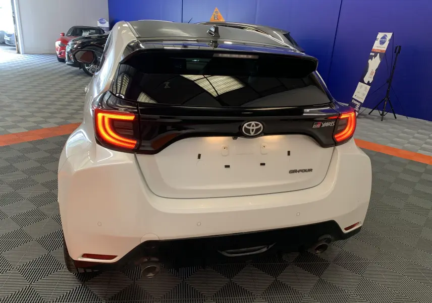 Vue arrière d'une Toyota Yaris GR blanche métal avec feux arrière allumés et toit noir dans un showroom.