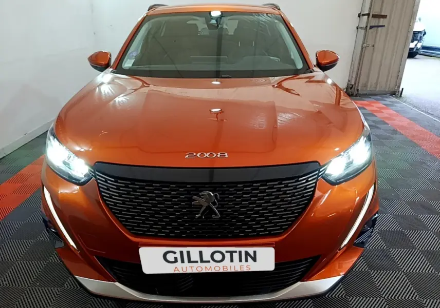 Vue avant d'un Peugeot 2008 orange avec phares LED allumés et calandre noire distinctive dans un garage.