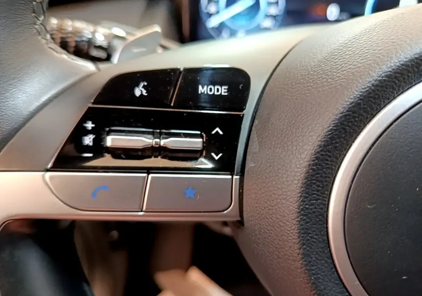 Gros plan sur les commandes gauche du volant cuir et alu du Hyundai Tucson bleu 2023, avec affichage digital en arrière-plan.