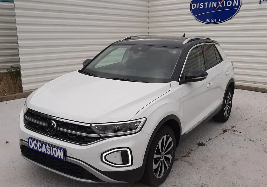 Volkswagen T-Roc blanc pur avec toit noir, vue 3/4 avant droit, stationné devant un mur blanc avec panneau Distinxion.