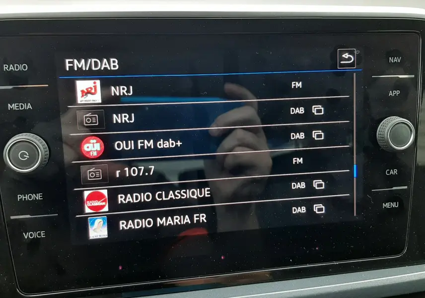 Vue rapprochée de l’écran tactile multimédia du Volkswagen T-Roc 2.0 TDI blanc avec toit noir, affichant les stations radio FM/DAB.