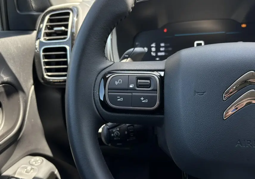 Gros plan sur le volant noir du Citroën C5 Aircross Hybrid 2025, avec commandes intégrées et aérateurs chromés à gauche.