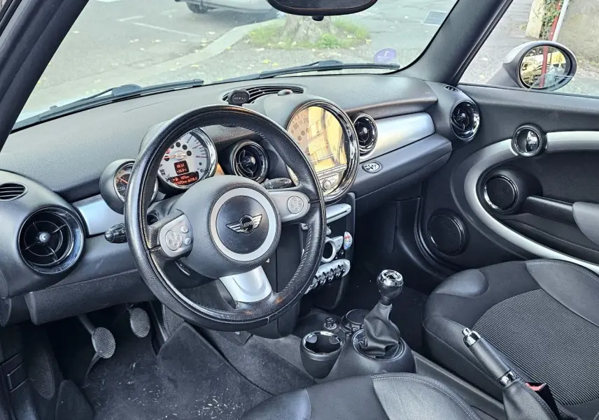 Vue intérieure côté conducteur d'une MINI Cabriolet R57 grise de 2010, avec volant cuir, tableau de bord rond et levier de vitesse manuel.