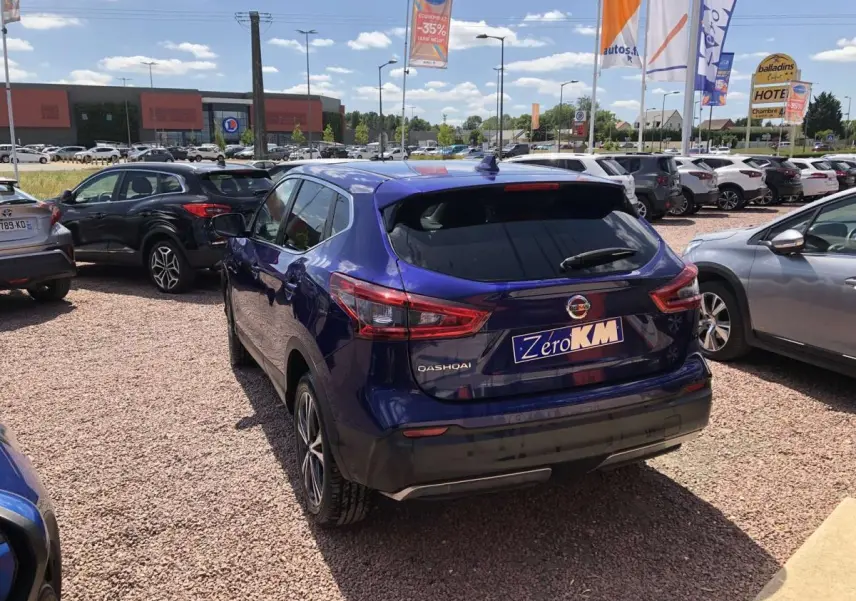 Vue 3/4 arrière d'un Nissan Qashqai bleu métallisé garé sur un parking extérieur sous un ciel ensoleillé.