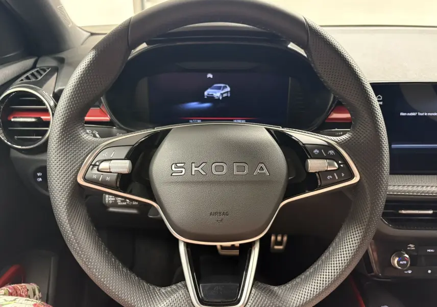 Vue rapprochée du volant perforé SKODA avec logo central, tableau de bord numérique et détails rouges du tableau de bord.