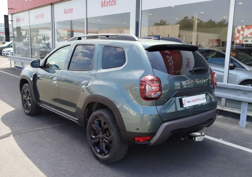Vue 3/4 arrière droite du Dacia Duster 2024 Vert Cèdre métallisé avec jantes noires et attelage visible.