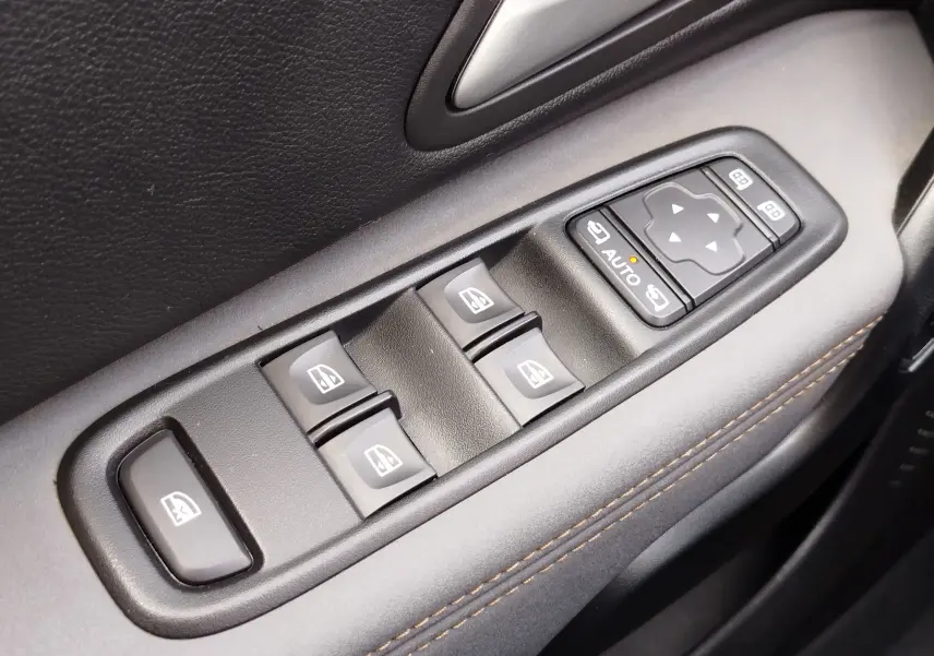 Gros plan sur les commandes des vitres électriques et des rétroviseurs côté conducteur d'un Dacia Duster 2024, garniture noire avec surpiqûres marron.
