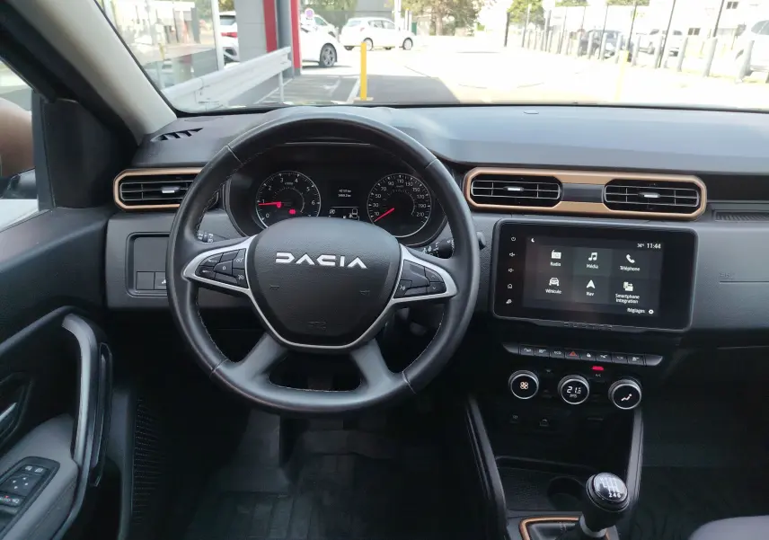 Vue intérieure du tableau de bord du Dacia Duster 2024, avec volant, écran tactile et levier de vitesses manuel.