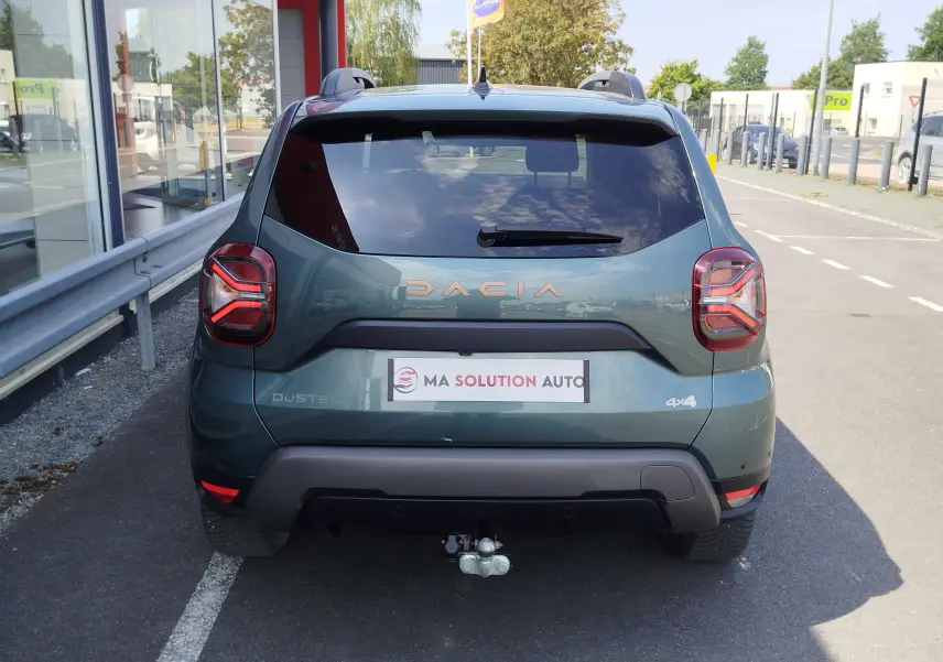 Vue arrière d'un Dacia Duster 2024 Vert Cèdre métallisé avec logo DACIA et attelage visible en stationnement.
