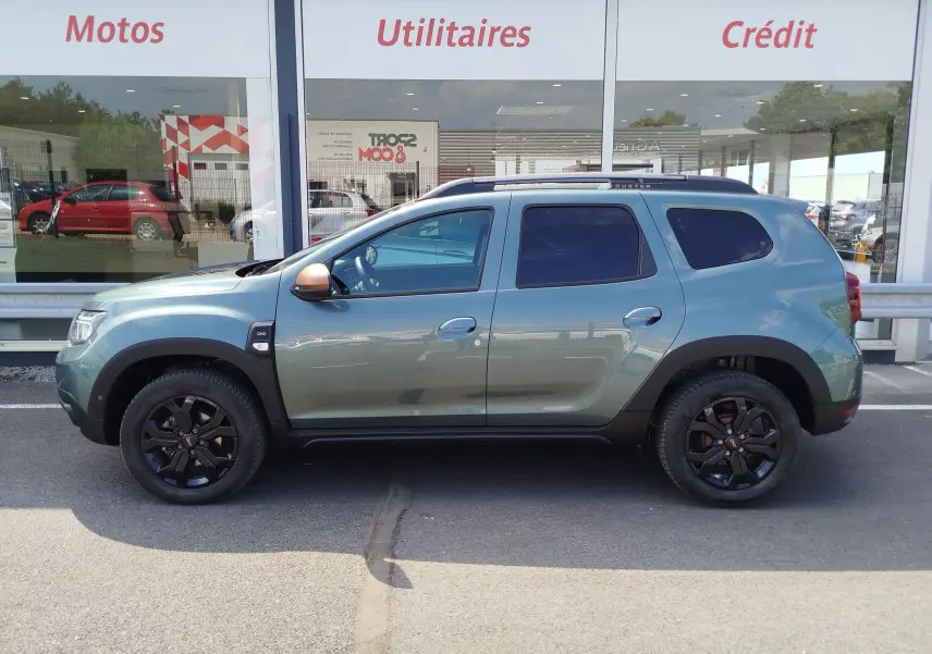 Vue de profil côté gauche du Dacia Duster 2024 Vert Cèdre métallisé avec jantes noires et barres de toit.