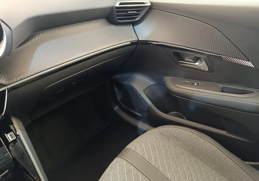 Vue intérieure côté passager de la Peugeot 208 électrique 2024 avec tableau de bord noir et siège tissu gris.