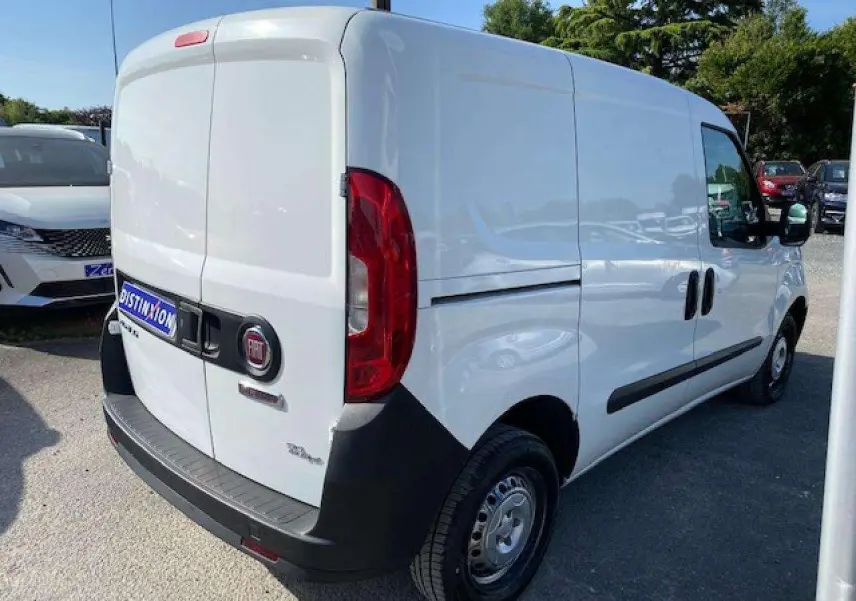 Fiat Doblo blanc vu en 3/4 arrière droit, fourgon tôlé avec pare-chocs noirs et porte latérale droite visible.