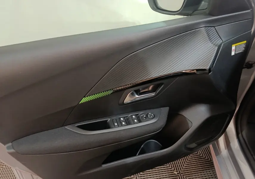 Vue intérieure côté gauche de la porte avant d’une Peugeot 208 électrique gris clair avec insert décoratif carbone et commandes fenêtres.