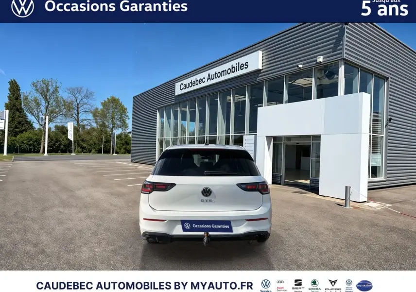 Vue arrière d'une Volkswagen Golf 1.5 eHybrid blanche avec attelage escamotable, stationnée devant un concessionnaire.