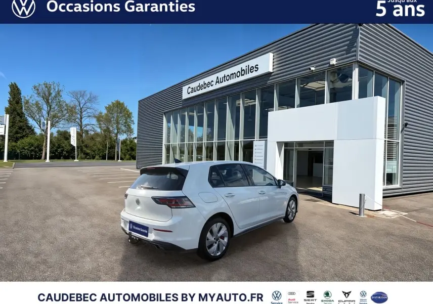 Vue 3/4 arrière droite d’une Volkswagen Golf 1.5 eHybrid blanche avec attelage escamotable, devant un bâtiment commercial.