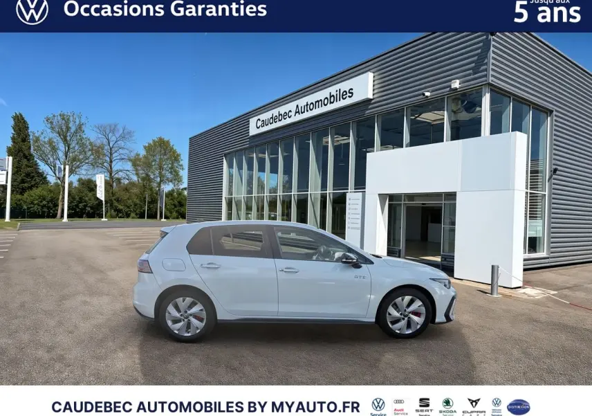 Vue latérale droite d'une Volkswagen Golf 1.5 eHybrid 272 DSG6 blanche stationnée devant un bâtiment commercial moderne.