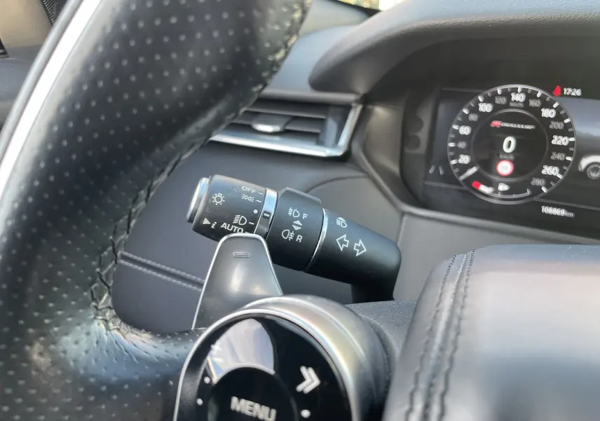 Gros plan sur le volant en cuir perforé et la commande des phares du Range Rover Velar noir Santorini, tableau de bord digital visible.