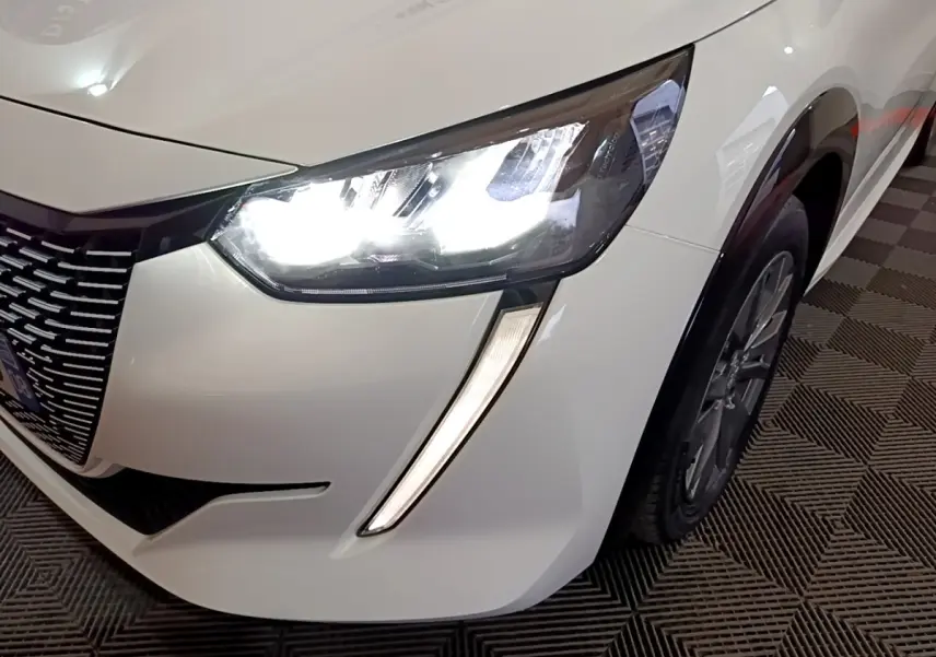 Gros plan sur l'avant droit blanc d'une Peugeot 208 électrique 2021, mettant en valeur le phare LED allumé et la calandre.