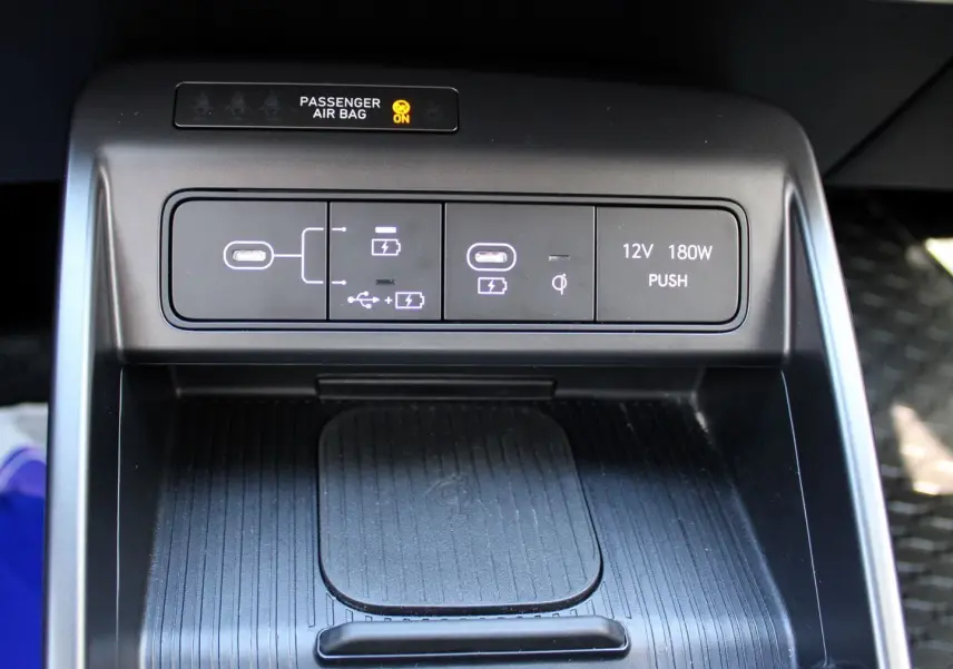 Gros plan sur la console centrale noire du Hyundai Kona 2024, montrant les ports USB, prise 12V et zone de recharge sans fil.