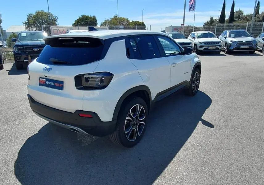 Jeep Avenger blanc en 3/4 arrière droit, avec feux arrière sombres et jantes alliage distinctives, sur parking extérieur.