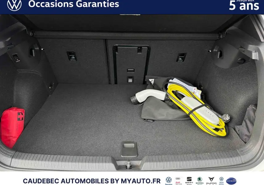 Coffre ouvert d’une Volkswagen Golf blanche avec câble de recharge électrique et trousse de secours rouge visible.