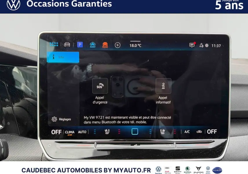 Vue intérieure centrée sur l'écran tactile du tableau de bord de la Volkswagen Golf blanche, affichant les commandes d'urgence et climatisation.