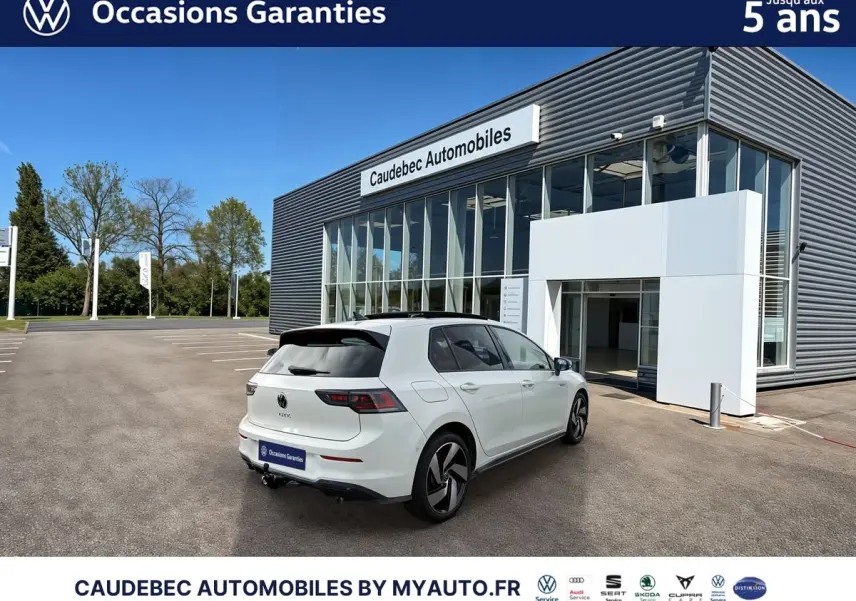 Vue 3/4 arrière droite d'une Volkswagen Golf 1.5 eHybrid blanche nacrée, stationnée devant un bâtiment moderne.