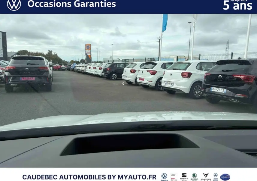 Vue depuis l’intérieur d’une Volkswagen Golf blanche, capot visible, sur un parking avec plusieurs voitures blanches et noires alignées.