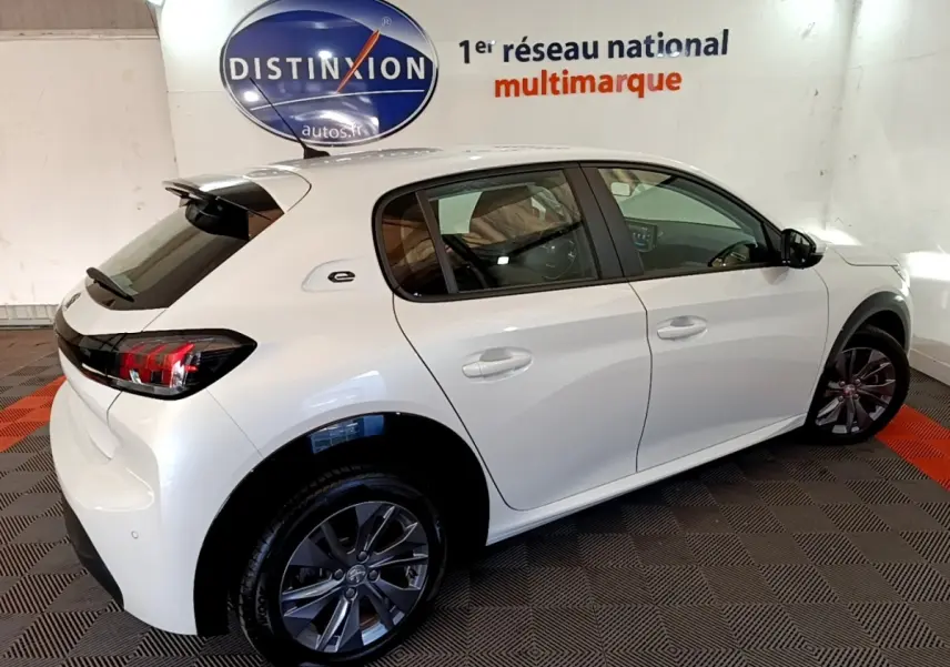 Vue 3/4 arrière droite d'une Peugeot 208 électrique Active Business blanche en intérieur, avec jantes foncées et logo "e" sur l'aile arrière.