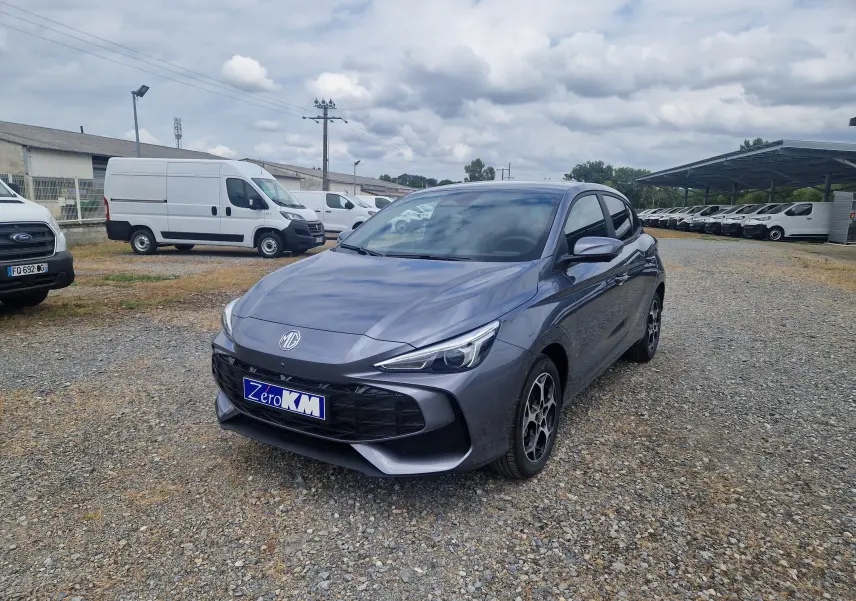 MG3 1.5L HYBRID+ 195CH Luxury en gris Hampstead, vue 3/4 avant sur un terrain gravillonné avec plusieurs utilitaires blancs en arrière-plan.