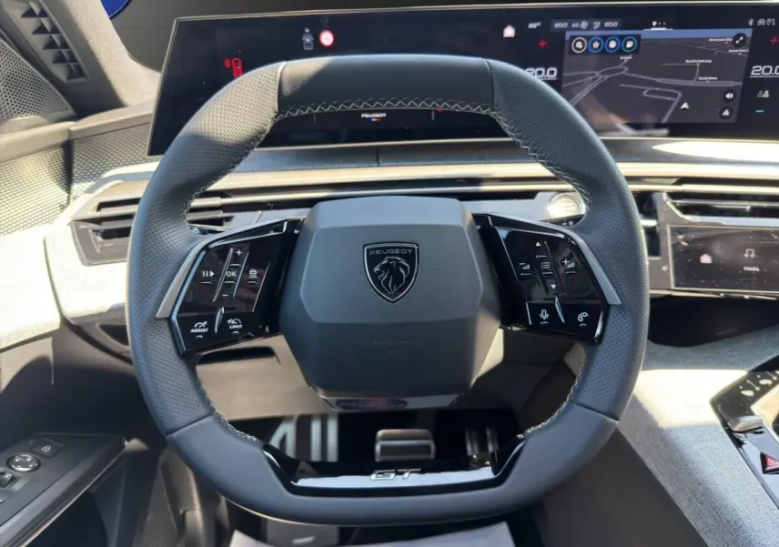 Vue rapprochée du volant cuir perforé noir avec logo Peugeot et inscription GT, tableau de bord numérique en arrière-plan.