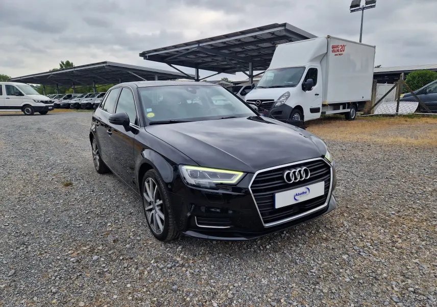 Audi A3 Sportback noire vue 3/4 avant droit sur un sol gravillonné avec des véhicules utilitaires en arrière-plan.